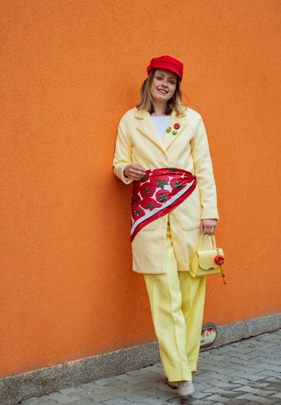 Mujer con abrigo y pantalones amarillos, gorra roja, sosteniendo un bolso amarillo con decoración de tomate, de pie frente a una pared naranja.