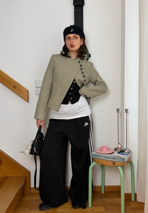 Joven mujer con boina negra, chaqueta beige, pantalones anchos negros, sosteniendo bolso negro, de pie en el interior cerca de unas escaleras de madera y una mesa pequeña con libros y velas.