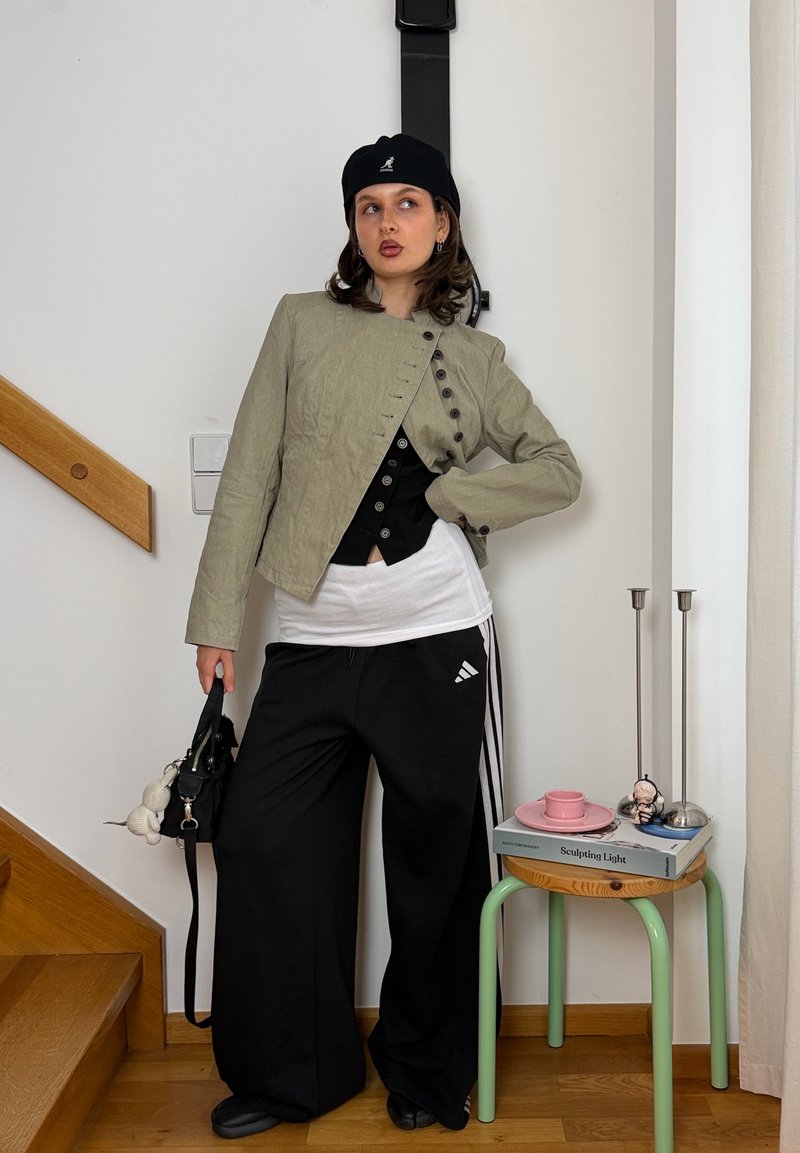 Joven mujer con boina negra, chaqueta beige, pantalones anchos negros, sosteniendo bolso negro, de pie en el interior cerca de unas escaleras de madera y una mesa pequeña con libros y velas.