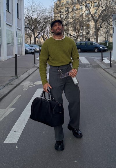 Hombre con suéter verde camina por una calle de la ciudad llevando una bolsa negra, sosteniendo gafas de sol y una botella de agua, con árboles sin hojas y edificios al fondo.