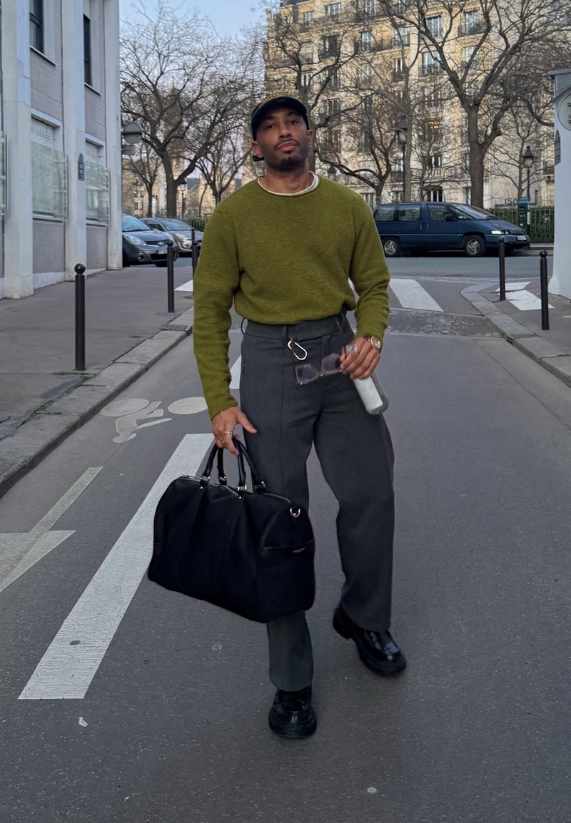 Homme en pull vert marchant dans une rue de la ville, portant un sac noir, tenant des lunettes de soleil et une bouteille d'eau, avec des arbres sans feuilles et des bâtiments en arrière-plan.