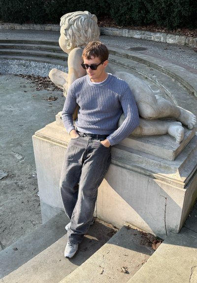 Suéter de punto gris con textura acanalada, combinado con jeans gris oscuro y zapatillas blancas. Sentado al lado de una escultura de piedra en un entorno de parque.