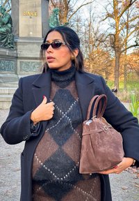 Brun semsket skinnveske med strammet topp og to håndtak. Modellen har på seg en mørk kåpe over en mønstret genser med diamantmotiv i nyanser av brun og grå.