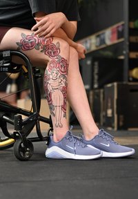 Tênis de atletismo cinza da Nike com solas brancas, combinados com uma tatuagem colorida de rosa e uma figura de estilo vintage na perna. Cadeira de rodas ao fundo.