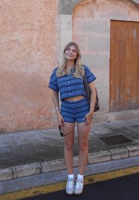 Dwuczęściowy zestaw w niebiesko-białe paski, krótki top z krótkim rękawem i szorty. Noszony z białymi skarpetkami i trampkami, trzymając okulary przeciwsłoneczne.