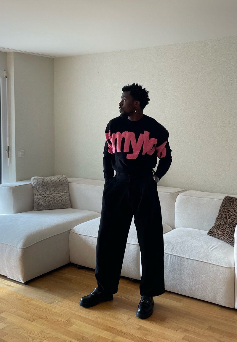 Homme avec des boucles d'oreilles portant un pull noir et rose et un pantalon large noir, debout dans un salon avec un canapé d'angle blanc et un sol en bois.