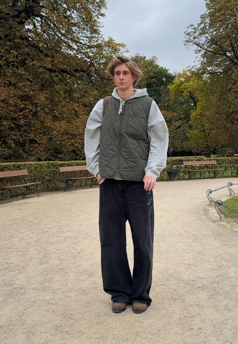 Ung person står på en grussti i en park, iført en grå hættetrøje, en olivengrøn quiltet vest, løse sorte jeans, brune sko og en rygsæk.