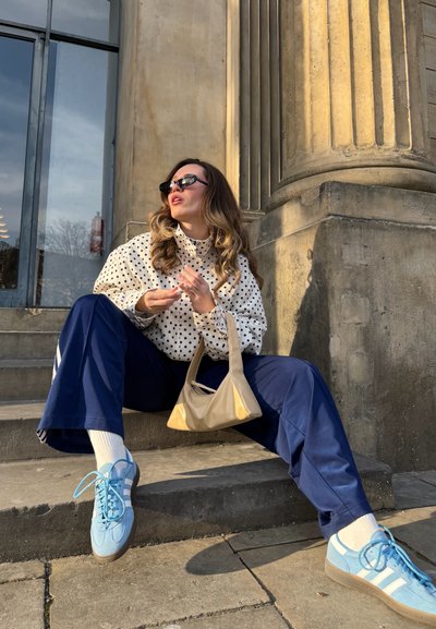 Mujer con blusa de lunares, pantalones azul marino, zapatillas azules y gafas de sol, sentada en unos escalones de piedra junto a una columna grande bajo la luz del sol, sosteniendo un bolso beige.