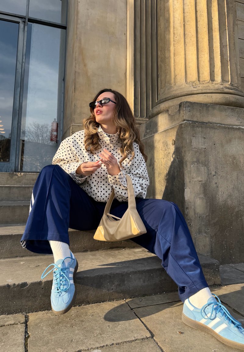 Femme portant un chemisier à pois, pantalon bleu marine, baskets bleues et lunettes de soleil, assise sur des marches en pierre près d'une grande colonne au soleil, tenant un sac à main beige.
