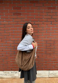 Νεαρή γυναίκα με μακριά σκούρα μαλλιά στέκεται μπροστά σε κόκκινο τοίχο από τούβλα, κρατώντας μεγάλη καφέ τσάντα ώμου από σουέντ, φορώντας στρωτά καθημερινά ρούχα.