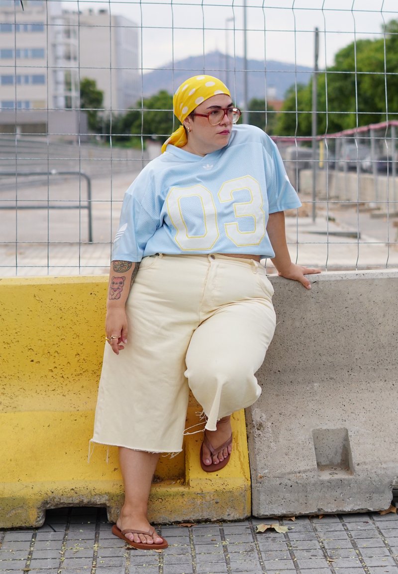Personne portant un foulard jaune à pois, de grandes lunettes, une chemise bleu clair avec le numéro 03, un pantalon crème et des sandales, assise sur une barrière jaune et grise.
