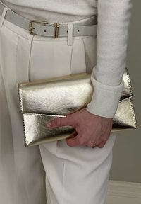 Main tenant une pochette enveloppe en métal doré, portant une ceinture blanche, un pull crème et un pantalon plissé crème sur un fond neutre.