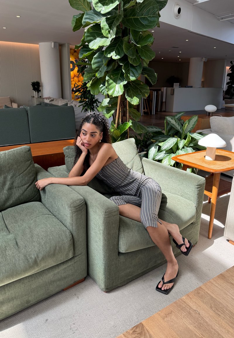 Femme en robe rayée, allongée sur des fauteuils verts dans un salon moderne avec de grandes plantes et des meubles en bois.