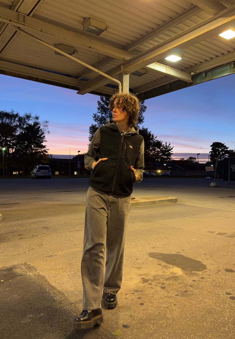 Schwarze Fleece-Weste über einem grauen Kapuzenpulli, graue weite Hose und schwarze Plateauschuhen. Außenbereich mit Sunset-Hintergrund.