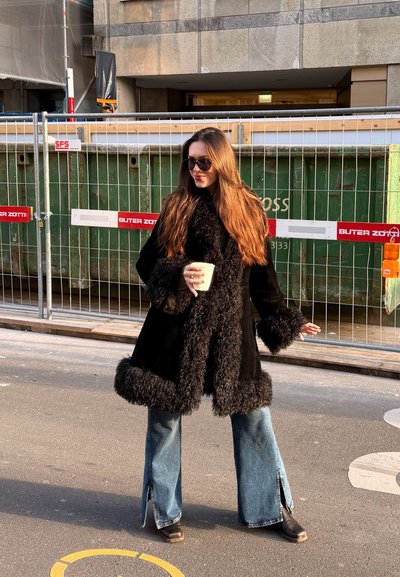Mujer con cabello largo y castaño, con abrigo negro con ribete de piel, gafas de sol, vaqueros azules, sosteniendo una taza de café, de pie en la calle cerca de una valla de construcción.