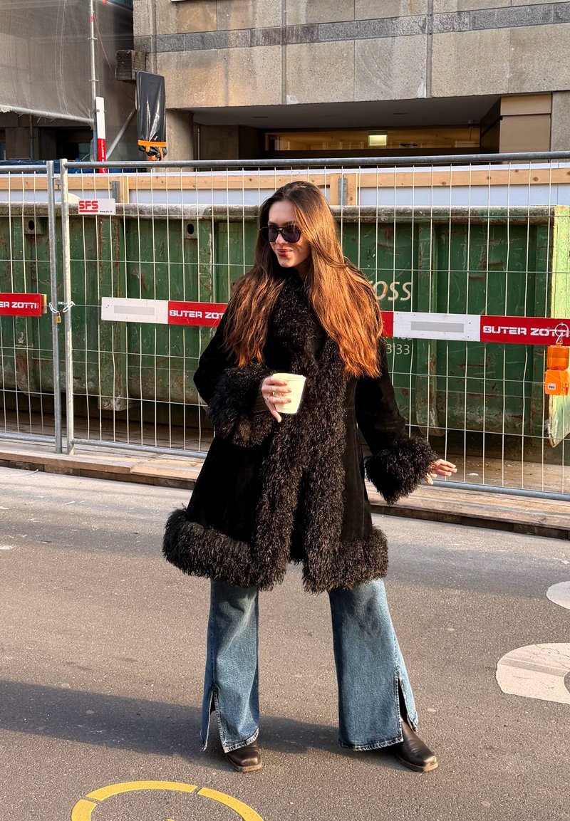 Femme aux longs cheveux bruns portant un manteau noir bordé de fourrure, des lunettes de soleil, un jean bleu, tenant une tasse de café, debout dans la rue près d'une clôture de chantier.