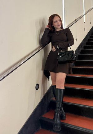 Joven mujer con vestido negro y botas hasta la rodilla está en las escaleras, apoyada en la barandilla, sosteniendo un bolso negro y tocándose el pelo.