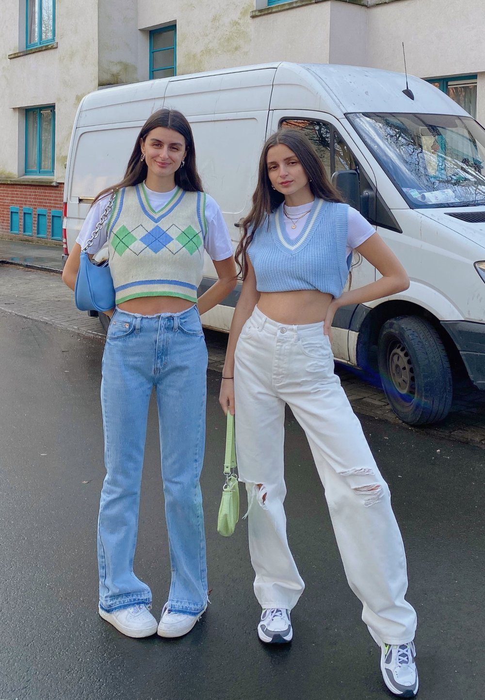 Dwie kobiety stoją na zewnątrz, jedna w kremowym kamizelce w wzór argyle i niebieskich dżinsach, druga w niebieskiej krótkiej kamizelce i podartych białych dżinsach.