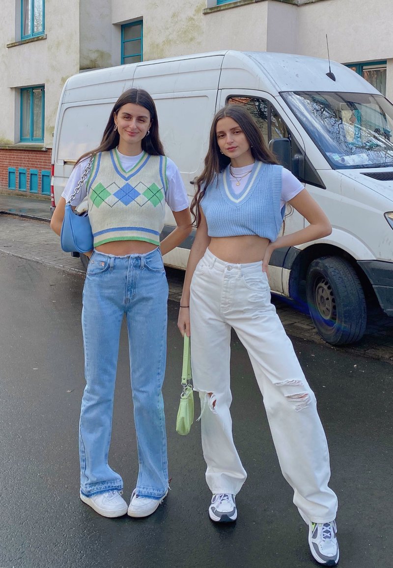 To kvinder står udendørs, den ene iført en cremet argyle vest og blå jeans, den anden i en blå cropped vest og slidte hvide jeans.