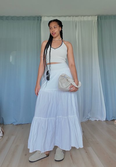 Falda maxi blanca con capas, top corto gris claro con escote halter, sosteniendo un bolso de mano beige. Pelo trenzado, calzado en tonos neutros.