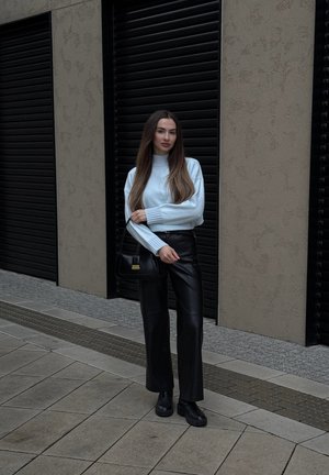 Mujer de pie sobre un pavimento de baldosas, vestida con un suéter azul claro, pantalones de cuero negros, botas negras y sosteniendo un bolso negro.