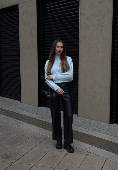 Mujer de pie sobre un pavimento de baldosas, vestida con un suéter azul claro, pantalones de cuero negros, botas negras y sosteniendo un bolso negro.