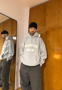 Sweat-shirt à capuche gris avec l'inscription "PROHIBITED BERLIN" en texte crème, associé à un pantalon cargo noir ample, debout à côté d'un miroir reflétant la tenue.