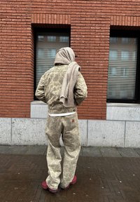 Μπεζ παντελόνι και εργασιακή ζακέτα με σχέδιο, σε χαλαρή εφαρμογή, συνδυασμένα με ένα ανοιχτό καφέ μαν τήλι, που είναι ριγμένο πάνω, και κόκκινα παπούτσια.