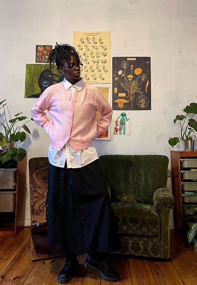 Cárdigan texturizado rosa sobre una camisa blanca de botones, combinado con una falda larga negra y botas negras. Sillón acolchado verde en el fondo.