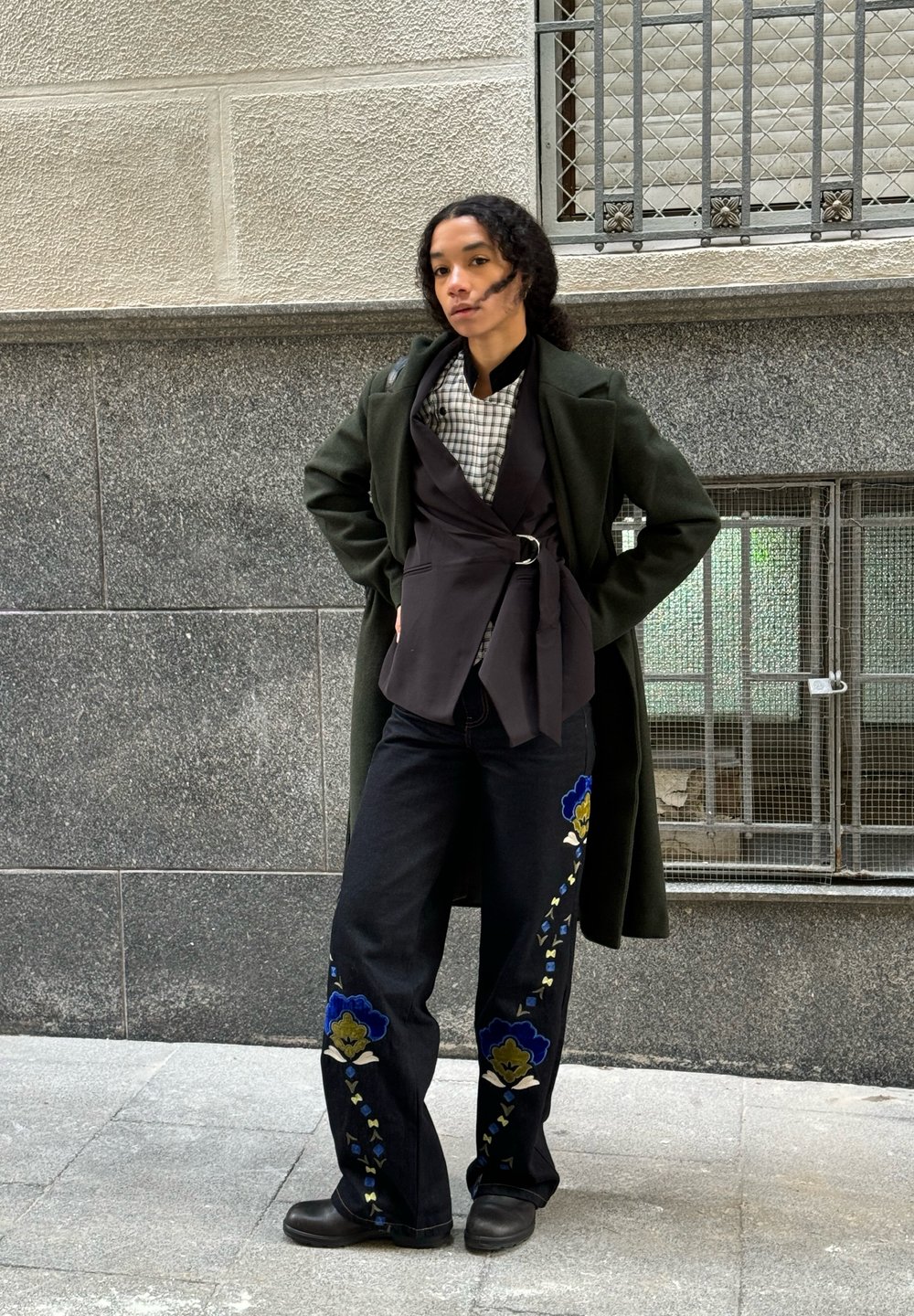 Tumši zaļš garš mētelis virs melna apakšveļas topa, pārbaudīta krekla un izšūtu melnu džinsu ar ziliem un dzelteniem ziedu rakstiem. Melni zābaki.