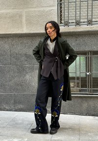 Tumši zaļš garš mētelis virs melna apakšveļas topa, pārbaudīta krekla un izšūtu melnu džinsu ar ziliem un dzelteniem ziedu rakstiem. Melni zābaki.