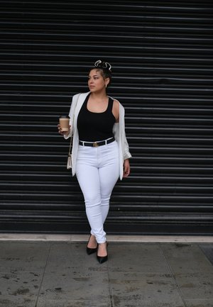 Top de tirantes negro, jeans blancos ajustados y una blazer de color claro. Sosteniendo una taza de café, con un cinturón estampado y zapatos negros de punta.