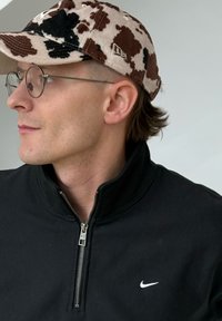 Άνδρας που φοράει στρογγυλά γυαλιά, καπέλο με τύπωμα καφέ και λευκής αγελάδας και μαύρο φερμουάρ μπουφάν Nike, κοιτάζοντας προς τα αριστερ ά σε σκηνικό με μονόχρωμο φόντο.