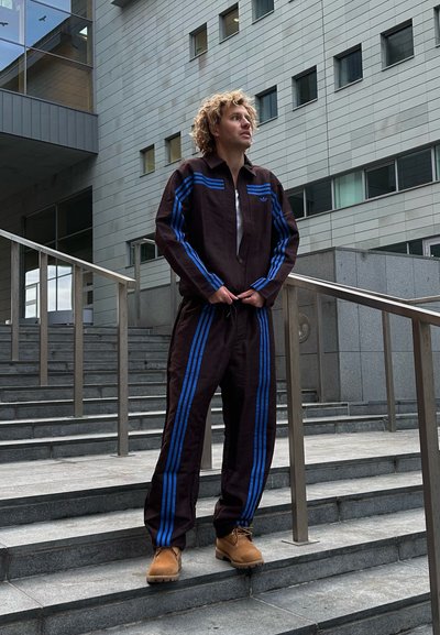 Chándal atlético marrón con rayas azules, que incluye una chaqueta con cremallera y pantalones a juego. La persona lleva botas beige y está de pie en unas escaleras.