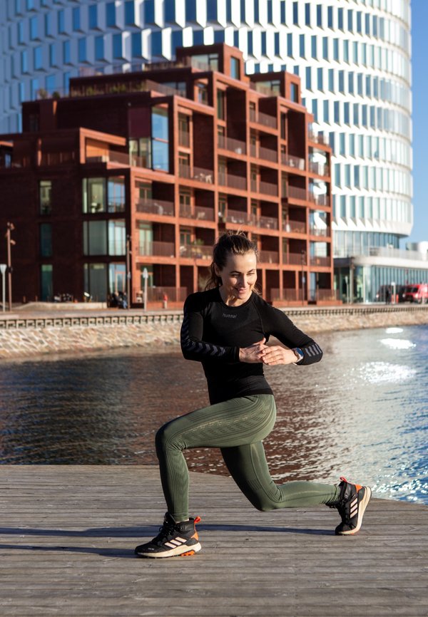 Svart langermet treningsgenser, mørkegrønne teksturerte tights og svarte joggesko med oransje detaljer. Vannfrontramme.