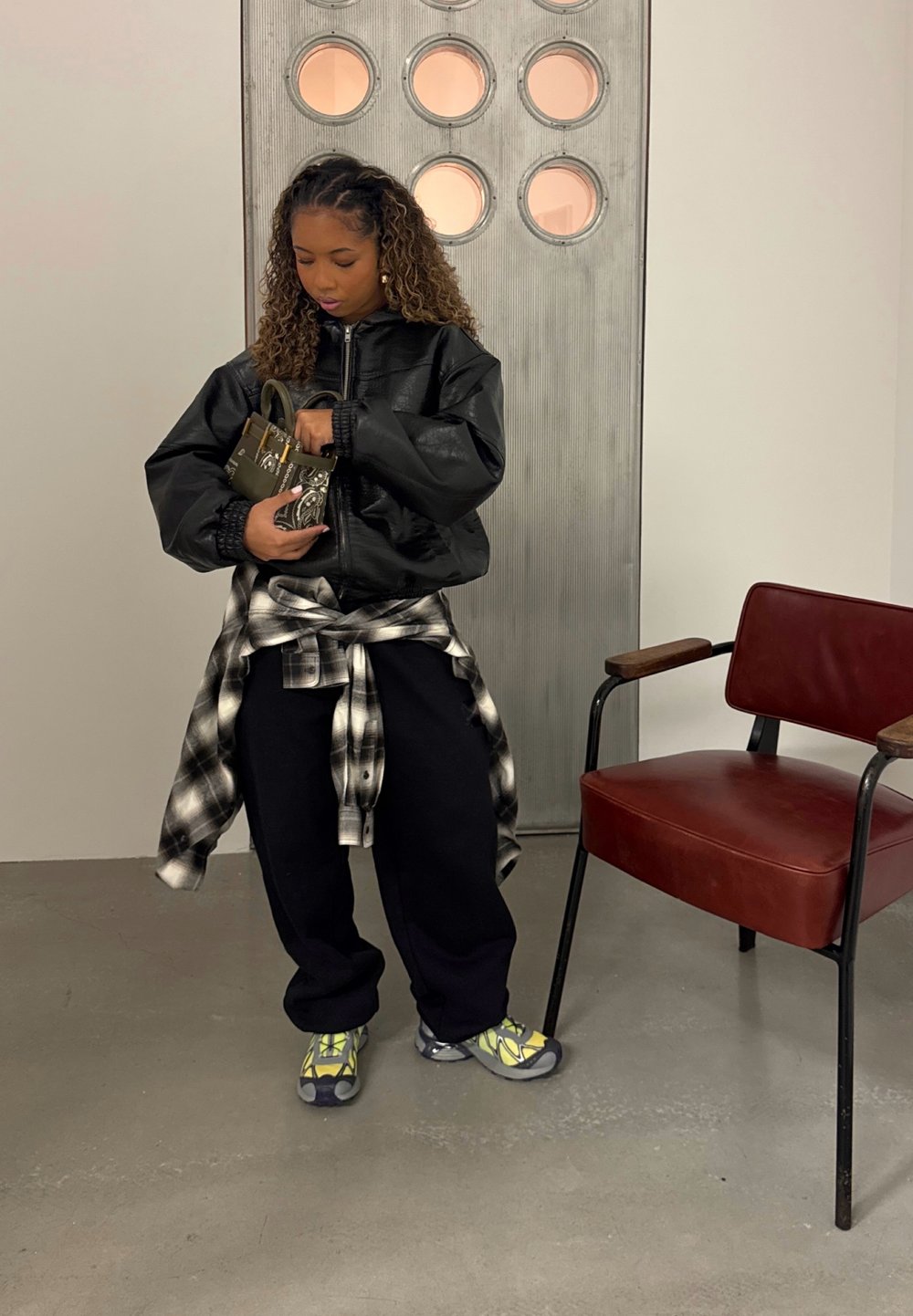 Jeune femme en tenue décontractée ajustant un petit sac à main à motifs, debout près d'une chaise rouge et d'une porte de style industriel avec des fenêtres rondes.