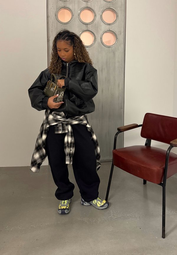 Ung kvinne i uformelt antrekk justerer en liten mønstret håndveske, står ved siden av en rød stol og en industriell dør med runde vinduer.