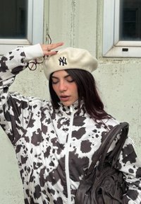 Jakke med kuprikk i svart og hvitt med høy krage, kremfarget beret med svart NY-logo, og en brun veske, foran en teksturert betongbakgrunn.