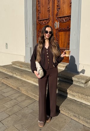 Mujer con cabello largo y ondulado, vistiendo un conjunto marrón oscuro y gafas de sol, sosteniendo un bolso blanco y un café con hielo, de pie en escalones de piedra junto a una puerta de madera.