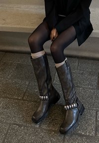 Hnědé kožené kozačky ke kolenům s hladkou texturou, mírným podpatkem a ozdobnými stříbrnými doplňky u kotníku. Stylizované s tmavými punčochami.