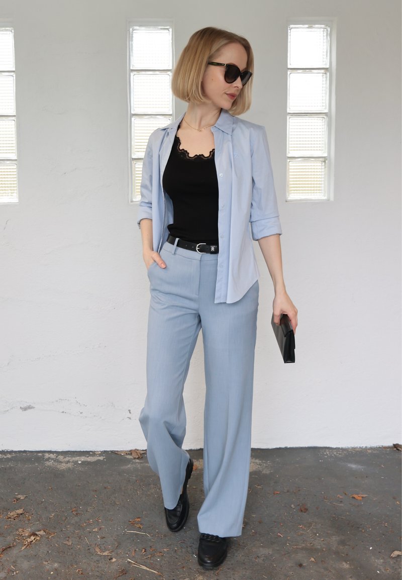 Femme portant un pantalon bleu clair et une chemise par-dessus un haut noir, lunettes de soleil noires, tenant une pochette noire, debout près d'un mur blanc avec des blocs de verre.