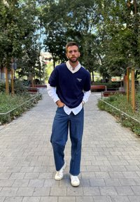 Jūras spēku džemperis virs baltas apkakles kreklas, brīvi zili džinsi un balti sporta apavi. Vides iezīme ir zaļa veģetācija un bruģēti ceļi.
