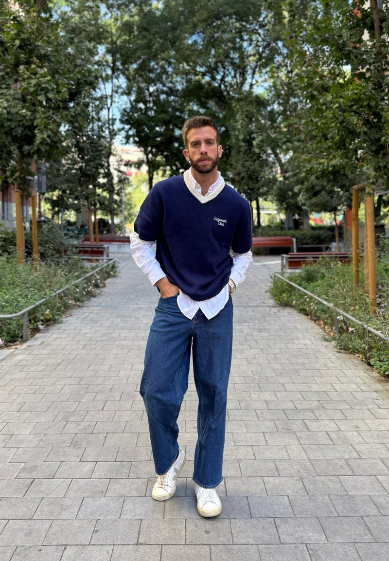 Maglione blu navy sopra una camicia bianca con colletto, jeans blu larghi e sneakers bianche. L'ambientazione presenta vegetazione e sentieri pavimentati.