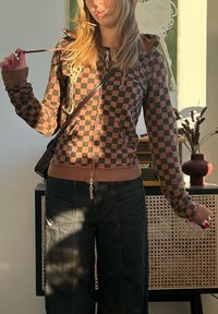 Personne portant un sweat à capuche à carreaux marron et noir et un jean noir, tenant le cordon du sweat à capuche, debout à l'intérieur près d'une armoire avec un vase et des fleurs séchées.