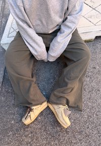 Personne assise sur le trottoir portant un sweat-shirt gris, un pantalon ample olive et des chaussures en daim beige avec des étiquettes latérales et des lacets décoratifs.