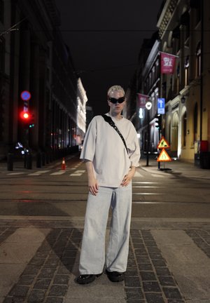 Camisa de gran tamaño gris claro, jeans de mezclilla de corte holgado claros, zapatos negros gruesos y gafas de sol oscuras. Entorno urbano nocturno con carteles de calles.
