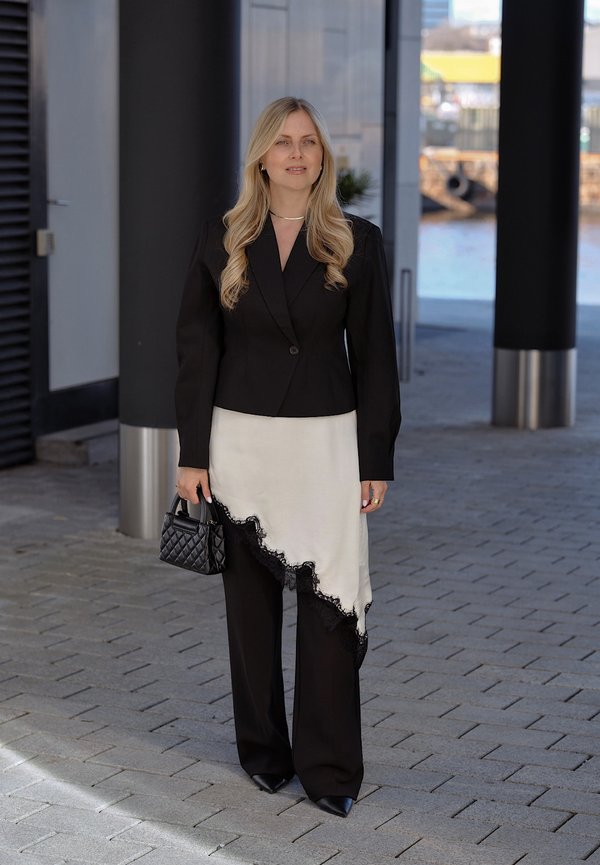 Femme blonde en blazer noir, sur-jupe blanche asymétrique avec dentelle noire, pantalon noir, tenant un sac à main noir matelassé, debout à l'extérieur.