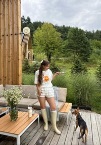 Hvit t-skjorte med oransje grafikk, volang hvite shorts, gule gummistøvler, tremøbler, glassvase med blomster, grønn utendørssetting.