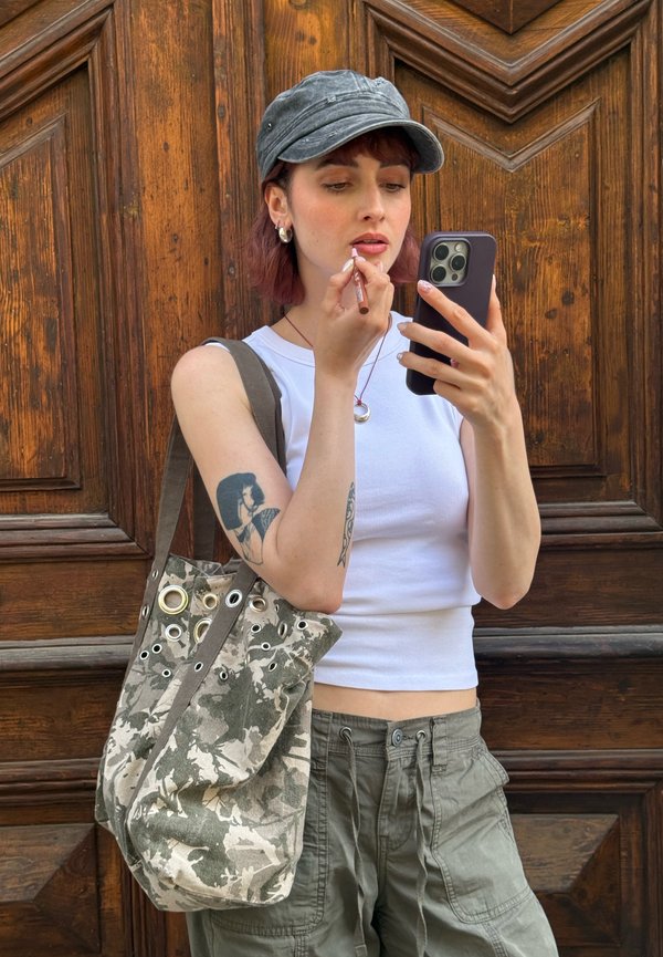 Jeune femme tatouée portant une casquette grise et un débardeur blanc applique du baume à lèvres en regardant son smartphone devant une porte en bois.