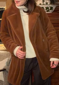Manteau en fausse fourrure marron avec un design à double boutonnage, grands revers et poches latérales. Porté sur un pull blanc et un pantalon foncé. Coupe décontractée.